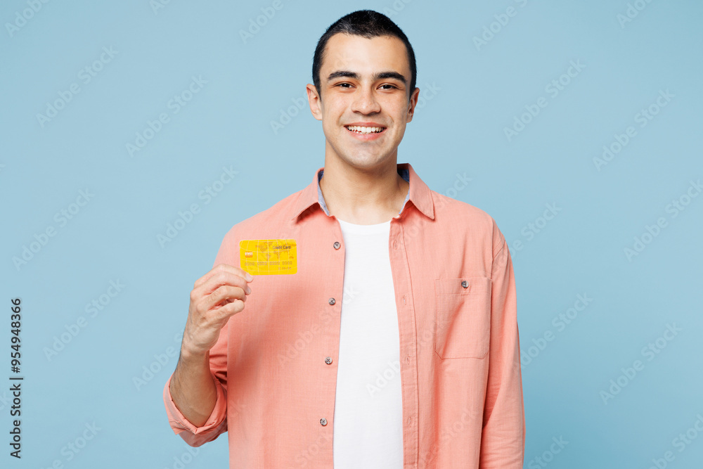 Young smiling happy middle eastern man wears pink shirt white t-shirt hold in hand mock up of credit bank card isolated on plain pastel light blue cyan background studio portrait. Lifestyle concept. Young smiling happy middle eastern man wears pink shirt white t-shirt hold in hand mock up of credit bank card isolated on plain pastel light blue cyan background studio portrait. Lifestyle concept.