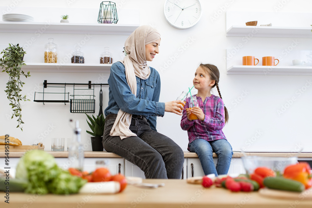 Woman in hijab sharing quality time with child surrounded by vegetables. Muslim mother and daughter drinking healthy juice together in modern kitchen setting. Family bonding over healthy lifestyle.