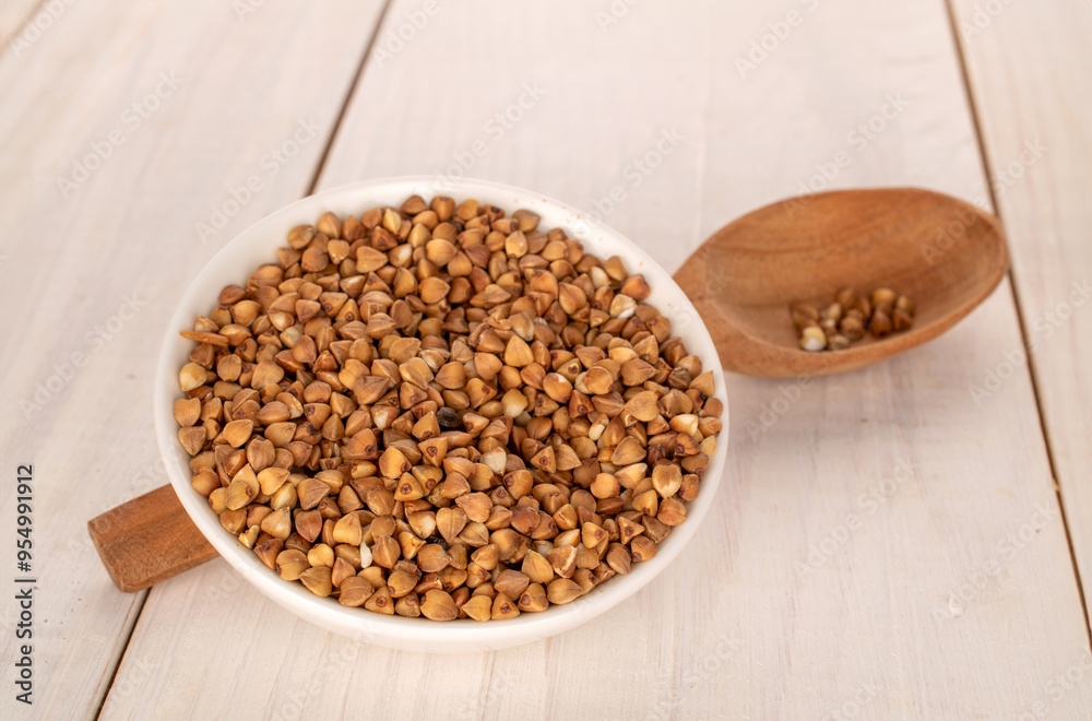 Raw buckwheat groats in a white ceramic dish with a wooden spoon on a wooden table, macro. Raw buckwheat groats in a white ceramic dish with a wooden spoon on a wooden table, macro.