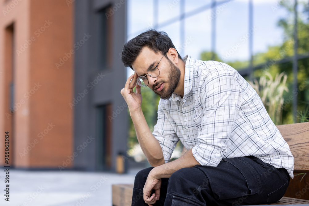 Tired young Indian man sitting on a bench outside an office building and holding his head with his hand, feeling a lot of pain and stress Tired young Indian man sitting on a bench outside an office building and holding his head with his hand, feeling a lot of pain and stress