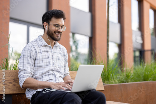 Tableau sur toile A smiling young Muslim man sits on a bench outside in the street wearing headpho