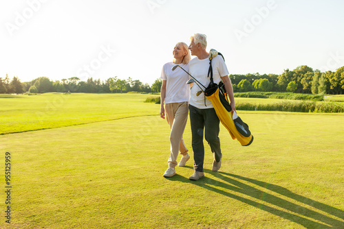 elderly senior couple in uniform holding golf clubs and walking on the background of golf course at sunset and talking, old man and woman walking and talking on golf game and looking at copy space