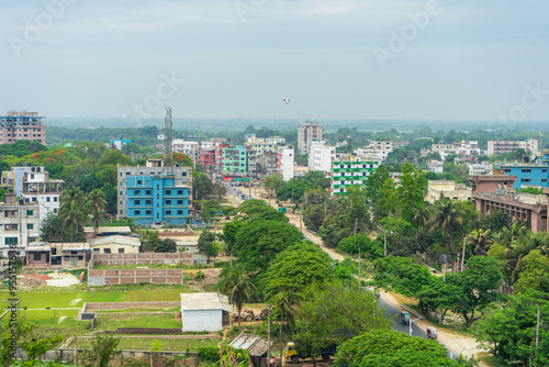 An aerial view of densely built-up area with various buildings of different sizes and colors, interspersed with patches of greenery under an overcast sky. Rajshahi, Bangladesh.