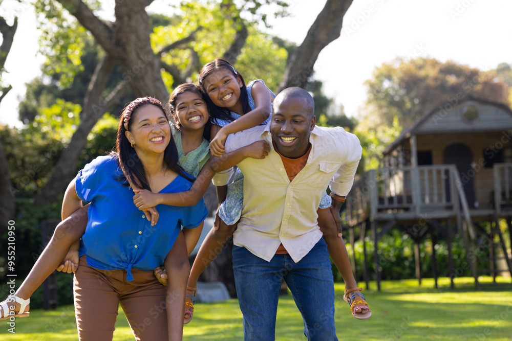 Smiling multiracial parents giving piggyback rides to happy daughters in backyard garden Smiling multiracial parents giving piggyback rides to happy daughters in backyard garden