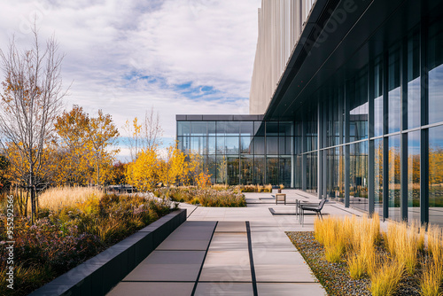 A contemporary building features a sleek glass exterior with a beautifully designed minimalist outdoor patio and lush surrounding autumn foliage under a clear sky.
