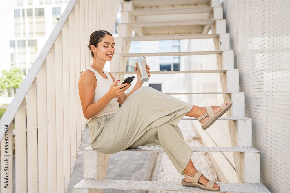 Fototapeta premium Young happy woman sit on stairs drink coffee and use mobile phone
