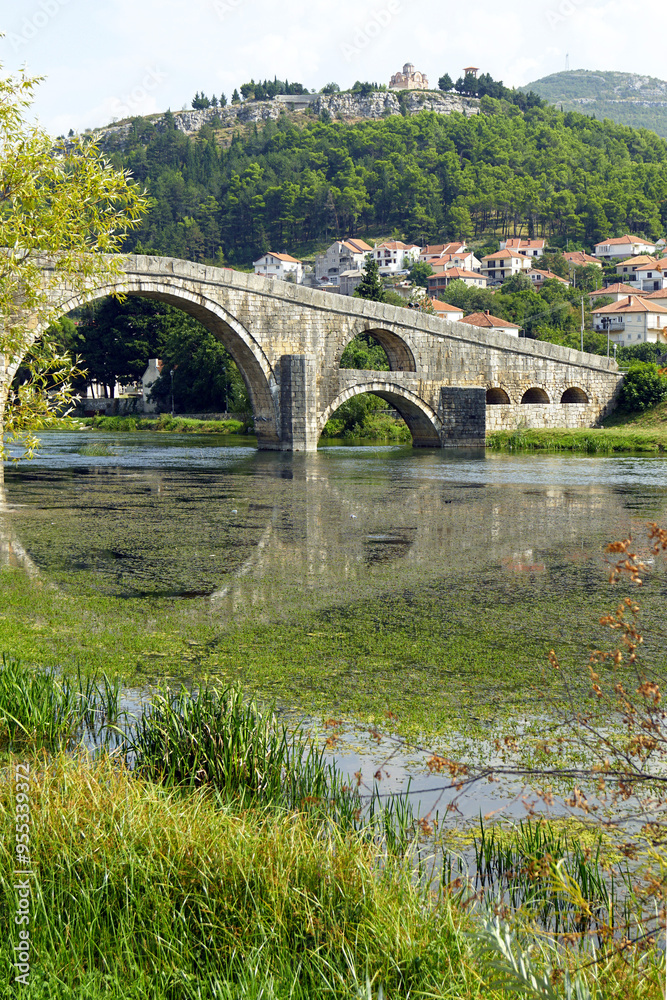 View from Trebinje, Bosnia and Herzegovina: grass on the bank, waters of the Trebišnjica River, arches of the Arslanagic stone bridge and Crkvice peak with the Hercegovacka Gračanica monastery. View from Trebinje, Bosnia and Herzegovina: grass on the bank, waters of the Trebišnjica River, arches of the Arslanagic stone bridge and Crkvice peak with the Hercegovacka Gračanica monastery.