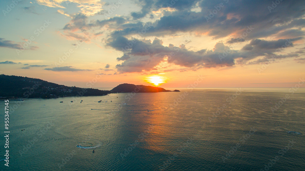 Naklejka premium A breathtaking aerial view of a coastal bay at sunset, with golden light reflecting on the calm sea and silhouetted hills in the distance. beautiful reflection of the golden sun at Patong Bay Phuket.