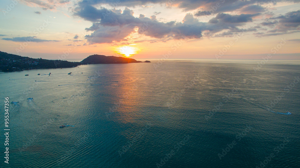 Obraz premium A breathtaking aerial view of a coastal bay at sunset, with golden light reflecting on the calm sea and silhouetted hills in the distance. beautiful reflection of the golden sun at Patong Bay Phuket.