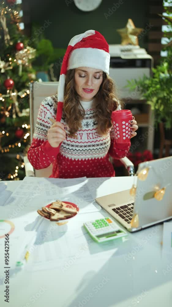 Christmas time. smiling modern in red Christmas sweater with traditional cookies, cup of tea and laptop singing in santa hat in modern green office with Christmas tree. Christmas time. smiling modern in red Christmas sweater with traditional cookies, cup of tea and laptop singing in santa hat in modern green office with Christmas tree.
