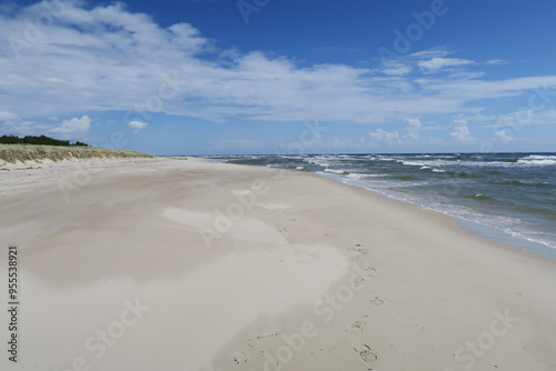 Wellen branden am Strand Sandhammaren in Ystad in der schwedischen Region Skane. Im Hintergrund sind weiße Schleierwolken zu sehen. Den Strand begrenzen Sanddünen mit Bewuchs. 