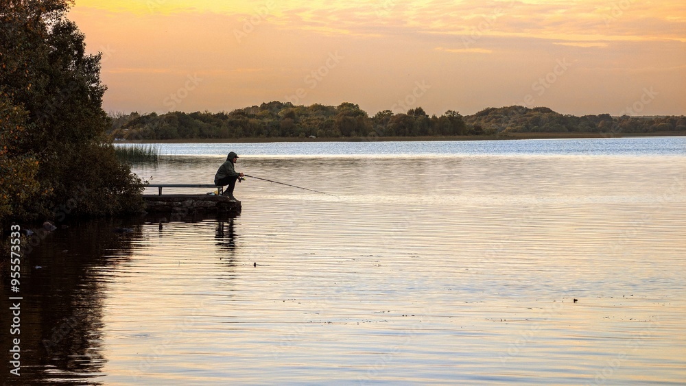 Silhouetted man fishing at sunset from pier at Ballyquirke lough, Galway, Ireland, sport and activities, catch and release, nature background