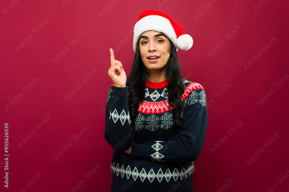 Young woman wearing a christmas sweater and santa hat holding up her index finger having an idea on red background Young woman wearing a christmas sweater and santa hat holding up her index finger having an idea on red background