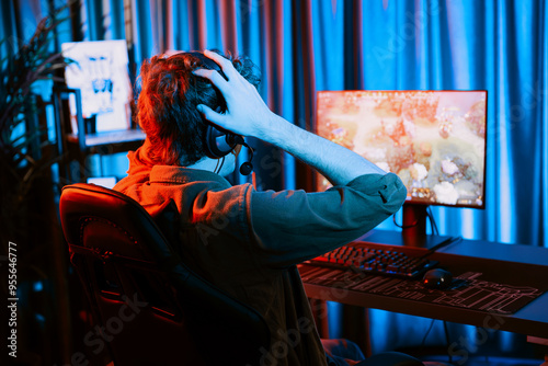 Host channel of stressful young gaming streamer getting to lose in fighting Moba at battle arena defeated multiplay team, wearing headphone on pc monitor with back side image at red neon room. Gusher. photo