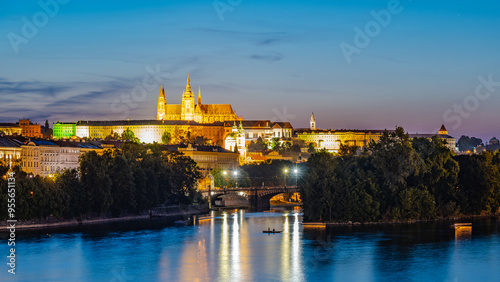 Prague Castle and the Vltava River are illuminated at night, with a small boat rowing across the water in the foreground. The reflections of the lights on the river create a mesmerizing scene.