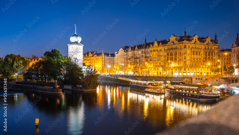 The Sitkov Water Tower stands tall along Masaryk Embankment in Prague, Czechia, as the sun sets and the city lights begin to illuminate the scene. The calm water reflects warm glow of the buildings. The Sitkov Water Tower stands tall along Masaryk Embankment in Prague, Czechia, as the sun sets and the city lights begin to illuminate the scene. The calm water reflects warm glow of the buildings.