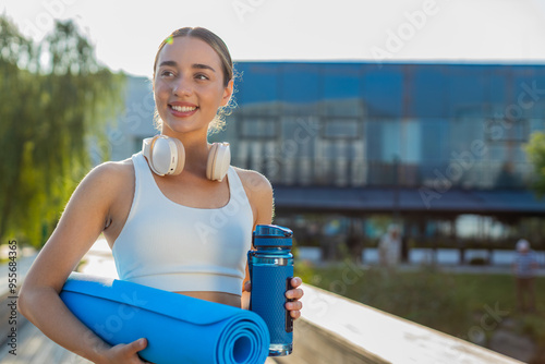 Young happy sportswoman with mat in her hands is heading to training outdoors in park. Smiling optimistic fit woman girl in activewear with headphones and water bottle goes to gymnastics training. photo