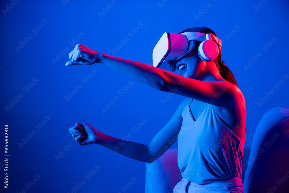Smart Female sitting on sofa surrounded by neon light wear VR headset connecting metaverse, futuristic cyberspace community technology. Elegant woman play car racing game of meta world. Hallucination. Smart Female sitting on sofa surrounded by neon light wear VR headset connecting metaverse, futuristic cyberspace community technology. Elegant woman play car racing game of meta world. Hallucination.
