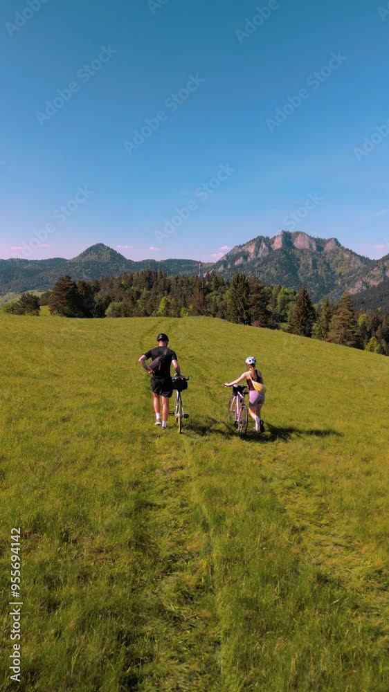 Trip to the mountains on bicycles. Two cyclists in the mountains. Two cyclists are pushing their bicycles. A man and a woman are riding their bicycles in a field with mountains in the background. Trip to the mountains on bicycles. Two cyclists in the mountains. Two cyclists are pushing their bicycles. A man and a woman are riding their bicycles in a field with mountains in the background.