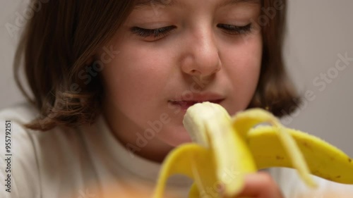 A close-up shot of a little girl taking a bite of a banana and eating it.​