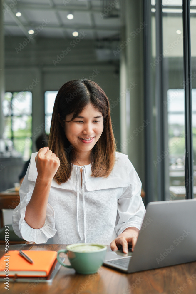 A young woman in a white blouse celebrates success while working on her laptop in a modern cafe, with a coffee cup and notebook on the table. A young woman in a white blouse celebrates success while working on her laptop in a modern cafe, with a coffee cup and notebook on the table.