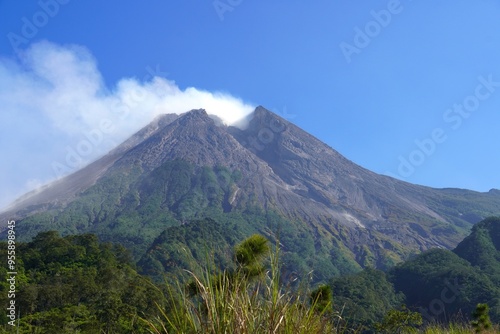 Mount Merapi in Yogyakarta, Indonesia, an active volcano with a split gray peak emitting white smoke, seen clearly against a bright blue sky