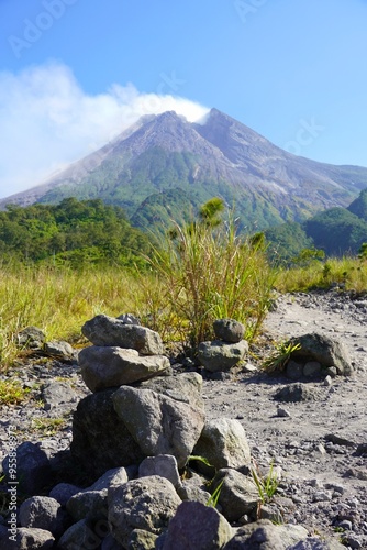 Mount Merapi in Yogyakarta, Indonesia, an active volcano with a split gray peak emitting white smoke, seen clearly against a bright blue sky
