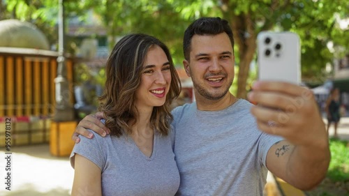 Couple taking a selfie outdoors in an urban park, with the man holding a smartphone while the woman smiles, enjoying a sunny day together in the city photo