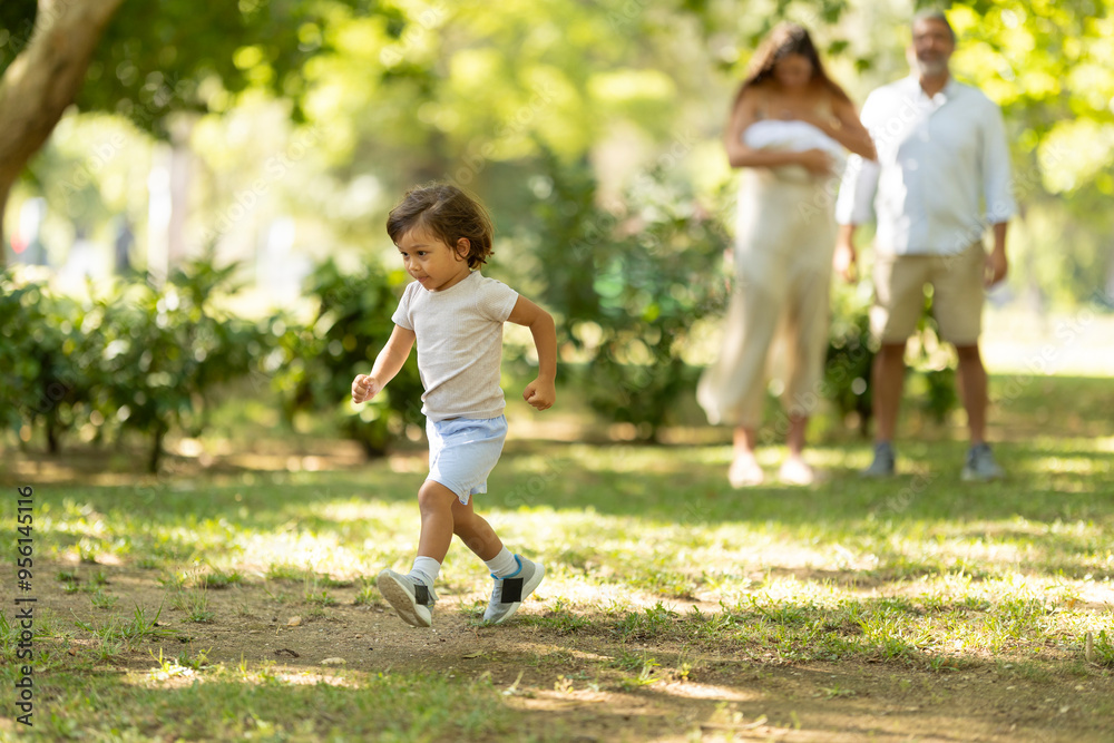 Little boy running happily in the park while parents watching in background Little boy running happily in the park while parents watching in background