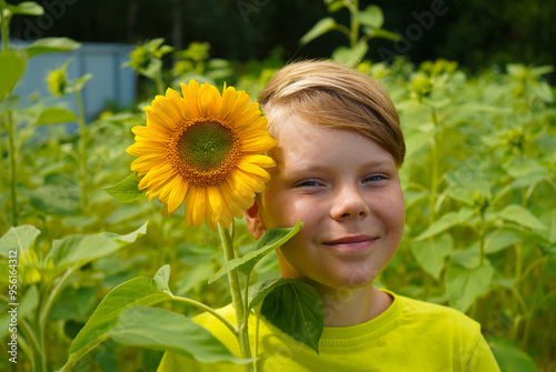 Carefree child with curly hair smiling and looking at camera while touching blooming flower on sunflower field in countryside