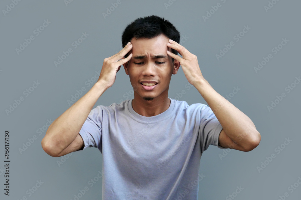 Young Asian man holding both sides of head with an expression of deep pain, looking tormented by migraine Young Asian man holding both sides of head with an expression of deep pain, looking tormented by migraine