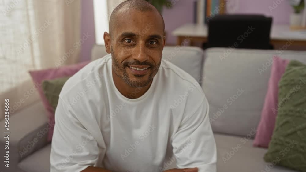 Handsome man sitting on a couch in an apartment living room, looking relaxed and smiling with indoor pillows and bookshelf in the background Handsome man sitting on a couch in an apartment living room, looking relaxed and smiling with indoor pillows and bookshelf in the background