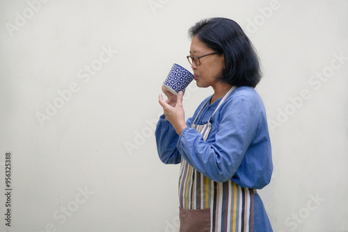 Asian woman standing side ways, wearing apron, sipping a cup of hot coffee photo