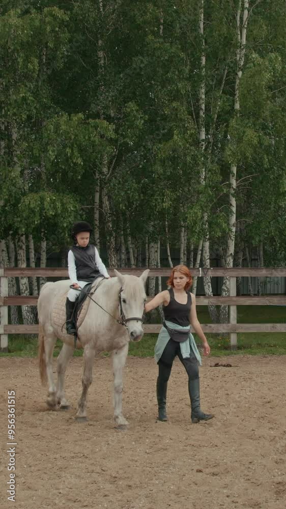 Vertical shot of female horse riding coach walking together with white horse with Caucasian boy with Down syndrome sitting in saddle on horseback in outdoor arena during therapeutic riding class Vertical shot of female horse riding coach walking together with white horse with Caucasian boy with Down syndrome sitting in saddle on horseback in outdoor arena during therapeutic riding class