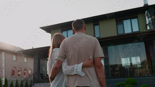 The couple is buying a big house. New large modern house. The family chooses a property, standing in front of building and discovering. Married couple looking at house, standing and hugging together 