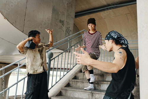 Group of Asian friends standing under overpass, with one wearing striped shirt and black beanie, and others gesturing expressively while engaged in lively conversation photo
