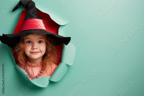 A young child wearing a witch's hat and pink attire joyfully peers through a colorful torn paper backdrop, capturing the essence of Halloween fun photo