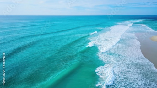an aerial view of a serene ocean scene with gentle waves rolling towards the shore, the water transitioning from deep blue to light green near the coast