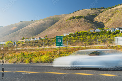 View of California 101 with a car driving under a bright, clear sky along a hilly road.