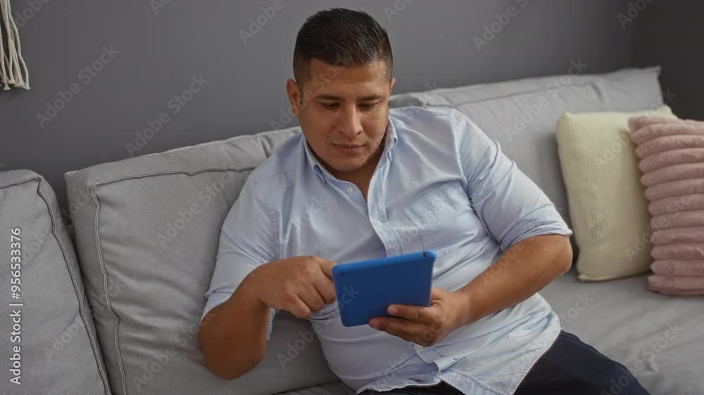 Young hispanic man using a tablet while relaxing on a couch in a living room at home Young hispanic man using a tablet while relaxing on a couch in a living room at home