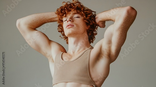 Nonbinary stylish person with curly red hair wearing beige crop top and low waist jeans showing his muscles on the arm pushing his lips together while standing against grey background : Generative AI