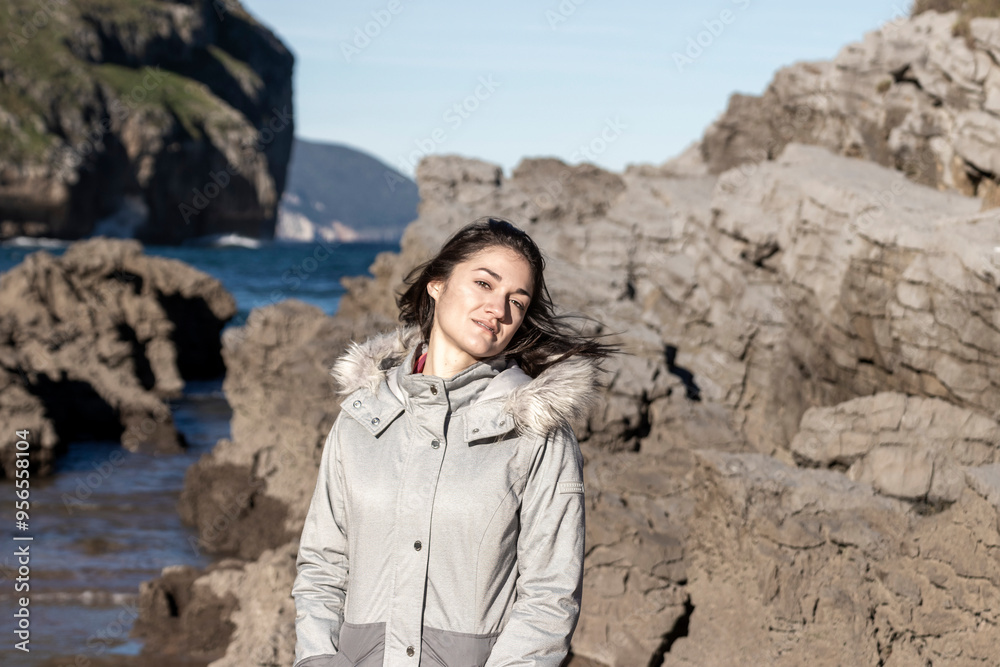 A woman is standing on a rocky beach with her hair blowing in the wind. She is wearing a grey jacket and smiling A woman is standing on a rocky beach with her hair blowing in the wind. She is wearing a grey jacket and smiling