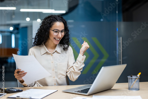 Wallpaper Mural Hispanic businesswoman celebrating achievement while working on laptop and holding document. Excitement and joy in modern office setting. Professional showcasing success and happiness in workplace. Torontodigital.ca