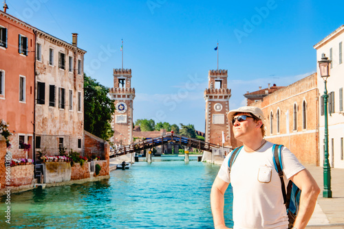 A cheerful traveler poses beside a picturesque canal with historic towers majestically in the background. Venice Arsenal complex of former shipyards and armaments are connected to each other.