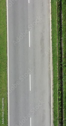 Vertical aerial view of a road with little traffic. View from sky of straight two-lane road. Direct above a paved transport road. White lane markings and gray pavement glow and reflecting in the sun