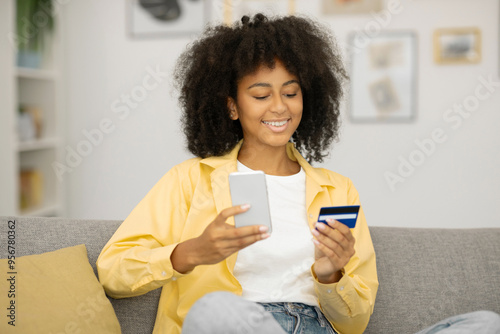 Happy African American lady shopping online with cellphone and credit card from home, sitting on sofa, enjoying e-commerce photo