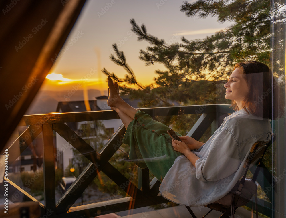 Young woman with smartphone greeting morning at dawn sitting on balcony overlooking mountains