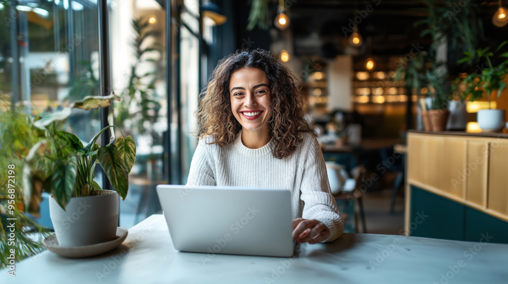 Cheerful woman with curly hair working on a laptop in a plant-filled modern cafe, smiling at the camera with large windows in the background. Cheerful woman with curly hair working on a laptop in a plant-filled modern cafe, smiling at the camera with large windows in the background.