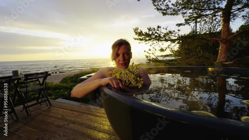 A woman enjoys a peaceful moment in an outdoor hot tub by the sea, holding a bouquet of flowers as the sun sets in the background. The golden light of the sunset casts a warm glow over the scene photo