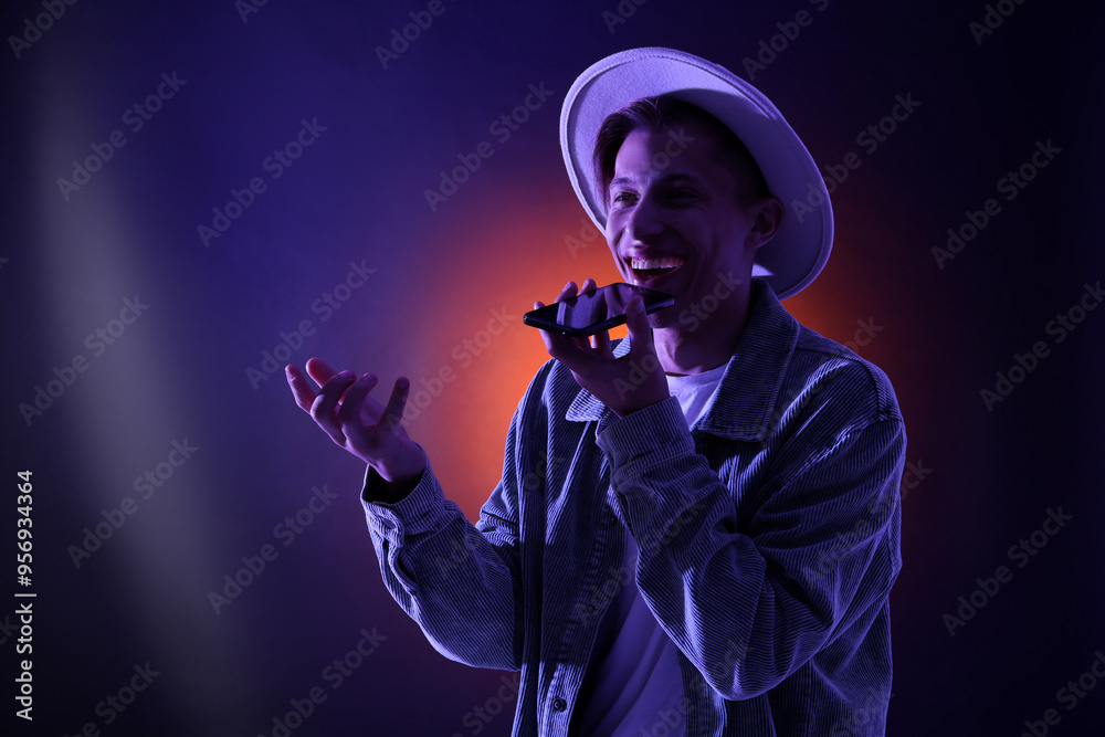 Young man recording voice message via smartphone on color background with neon lights, space for text Young man recording voice message via smartphone on color background with neon lights, space for text